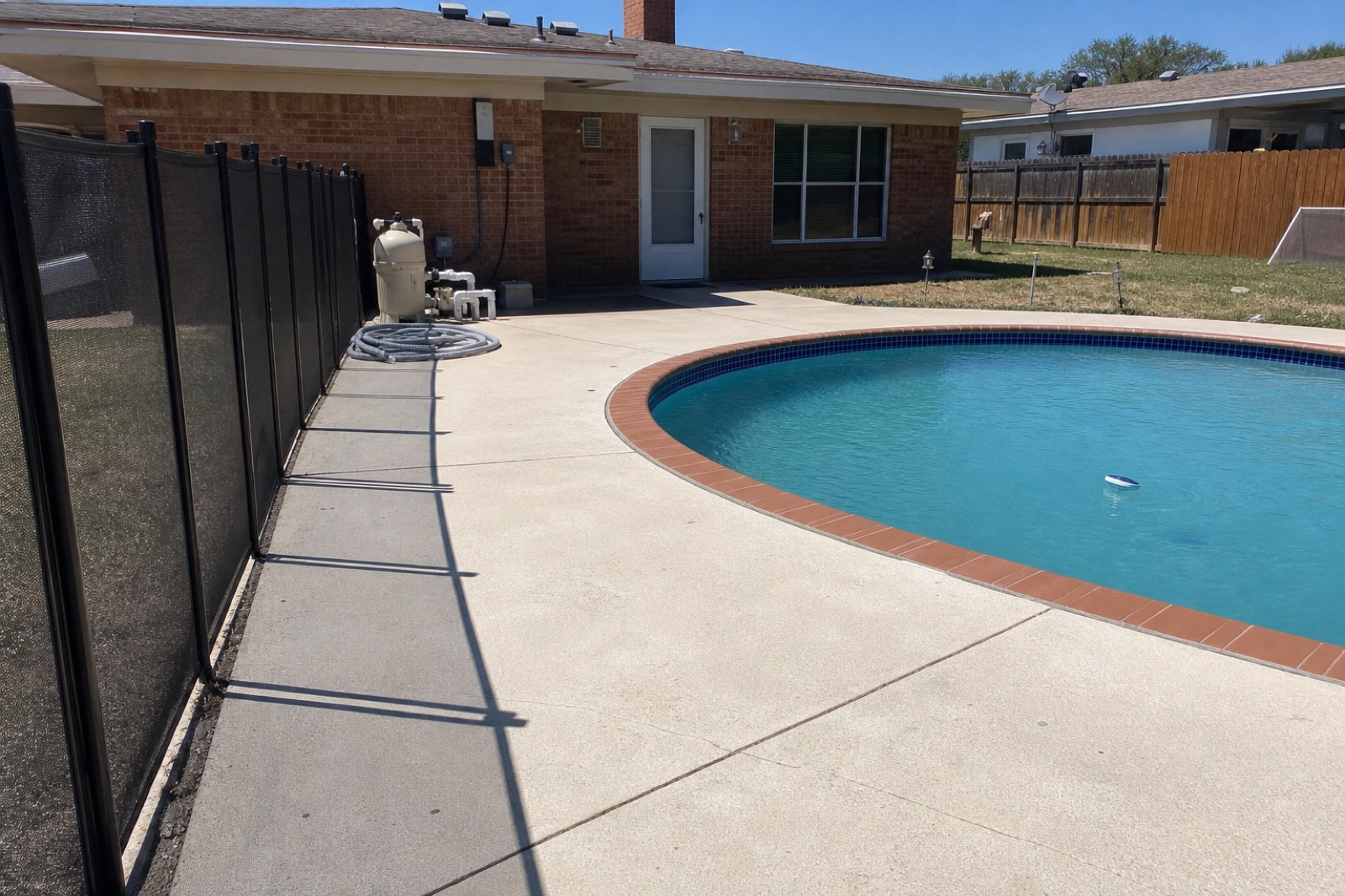A McKinney backyard with an in-ground swimming pool, brick ranch house and equipment pad visible behind a black mesh safety fence under harsh midday Texas sun.