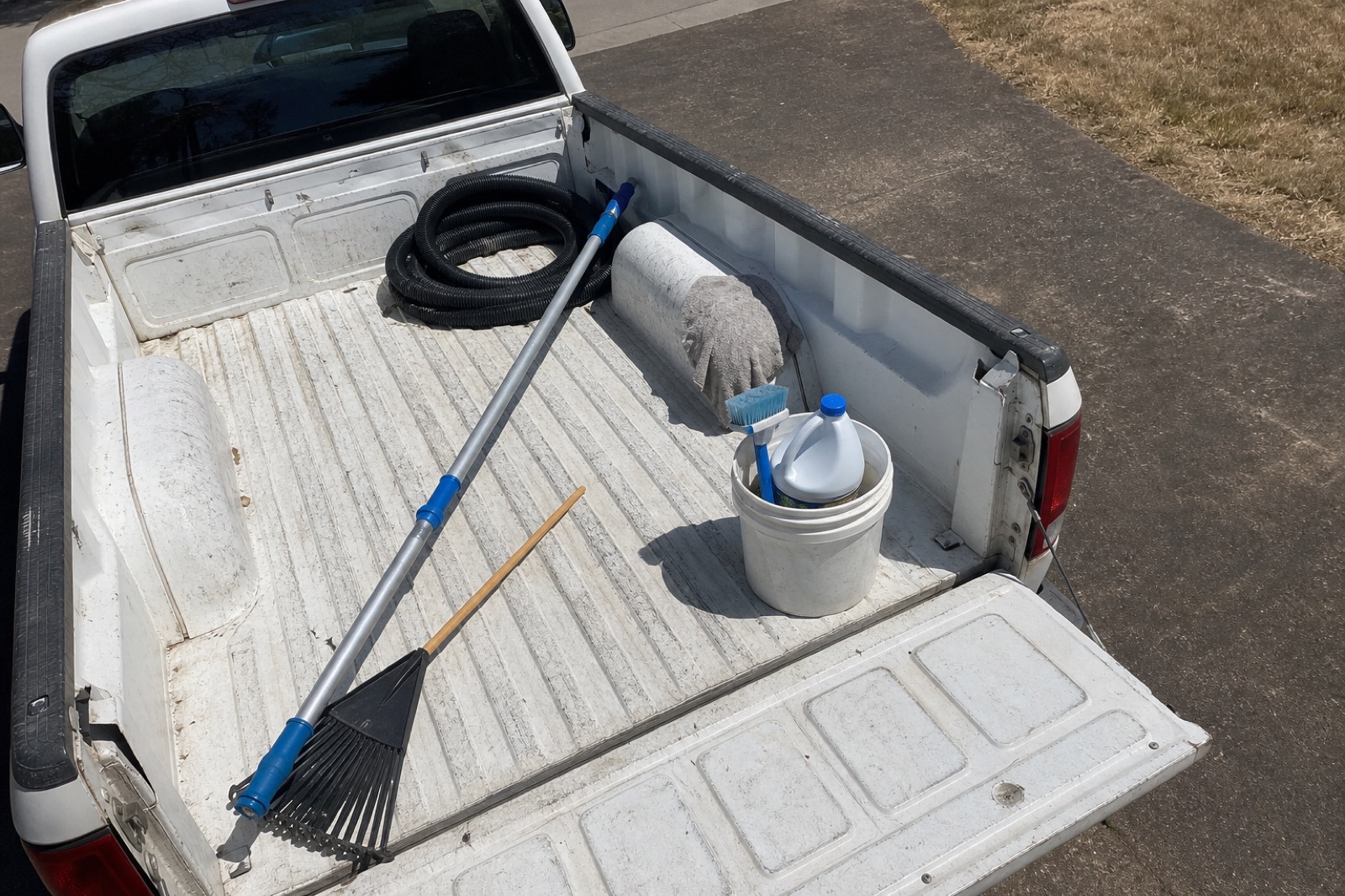 The open bed of a white work pickup truck in a McKinney driveway with a telescoping pool pole, blue leaf rake, coiled black backwash hose, a five-gallon bucket holding a tile brush and a bottle of muriatic acid, and a stained microfiber towel.