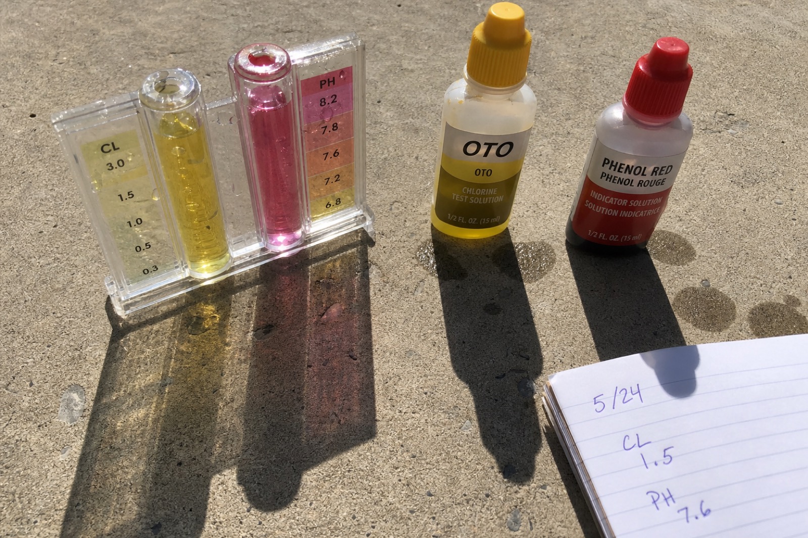 A pool water-testing comparator block with yellow chlorine and pink phenol-red sample wells beside reagent bottles, with a small notebook showing handwritten chlorine and pH numbers in ballpoint pen.